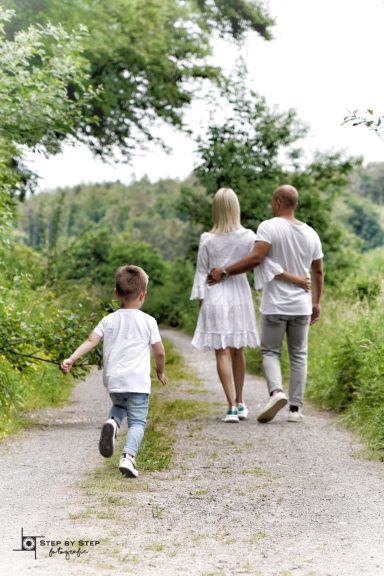 Eine Familie mit einem Kind spaziert auf einem Weg, umgeben von Bäumen und Grün.